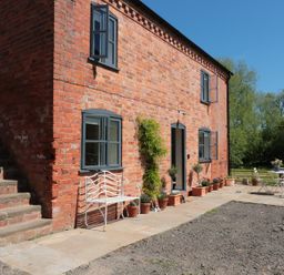 An exterior view of a brick building with open windows a white metal bench potted plants and a white table with chairs outside Granary 2 in Madley