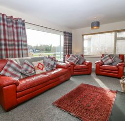 A living room with red leather sofa and chairs with plaid cushions a red rug on a grey carpet and large windows with blinds and curtains at Cae Glas in Pwllheli