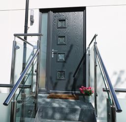 A black front door with glass panes and a metal railing with glass panels on stairs and a potted plant beside the door at An Drowes in Newquay