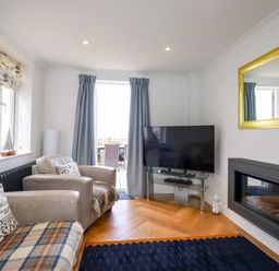 A living room with two armchairs a television on a glass stand a wall-mounted fireplace and a large mirror at Pink House in Mevagissey