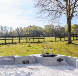 An outdoor hot tub with two glasses of champagne on the edge overlooking a grassy fenced field with trees at Pheasant Lodge in Wimborne Minster