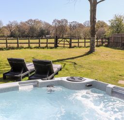 An outdoor hot tub with two lounge chairs on grass near a tree and a wooden fence at Pheasant Lodge in Wimborne Minster