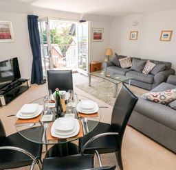 A living room with a glass dining table set for four black chairs two grey sofas a glass coffee table television and open balcony doors at 24 Rhodewood House in Saundersfoot