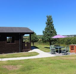 An outdoor seating area with chairs a table and umbrella next to a barbecue with a wooden cabin and trees in the background at Willow Lodge in Lanivet near Bodmin