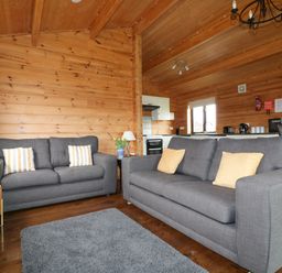 A living room with two grey sofas cushions a grey rug wooden walls and ceiling with a view into a kitchen at Holly Lodge in Lanivet near Bodmin