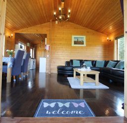 A wooden living room with a black sectional sofa and coffee table near a dining area with blue chairs at Hazel Lodge in Lanivet near Bodmin