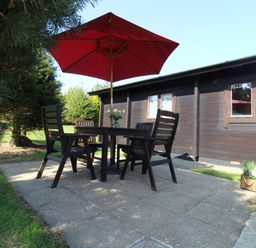 An outdoor patio with a table and four chairs under a red umbrella next to a wooden building with windows at Cherry Lodge Lanivet near Bodmin