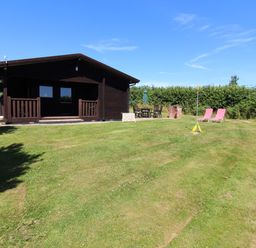 A wooden cabin with a porch on a lawn with chairs a table a grill and two pink lounge chairs at Maple Lodge in Lanivet near Bodmin