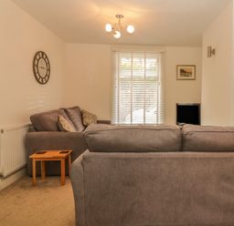 A living room with grey sofas, a wooden side table, a wall clock, and a window with blinds at Sunvale in Teignmouth