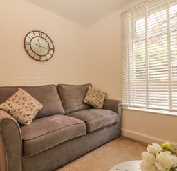 A room with a grey sofa with patterned cushions a wooden side table a wall clock white blinds on the window and flowers on a table at Sunvale in Teignmouth