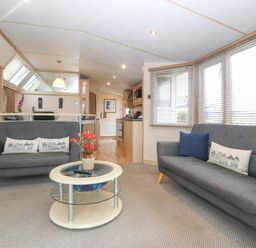 A living room with two gray sofas a round glass coffee table with flowers and a kitchen in the background at Coed Llai Lodge in Trearddur Bay
