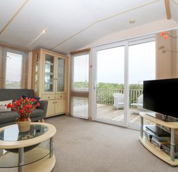 A living room with a sofa glass cabinet coffee table television and sliding doors leading to a wooden deck at Coed Llai Lodge in Trearddur Bay