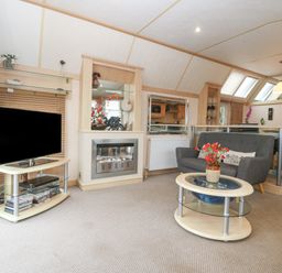 A living area with a gray sofa, a glass coffee table with a flower vase, a television on a stand, and a fireplace at Coed Llai Lodge in Trearddur Bay