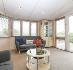A living room with sofas a round coffee table with flowers and a glass cabinet by windows at Coed Llai Lodge in Trearddur Bay