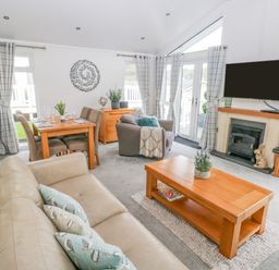 A living room with beige sofa, wooden coffee table with plant, dining table with chairs, armchair, wall-mounted TV above fireplace and patterned curtains at The Lookout in Stepaside