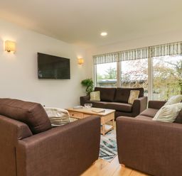 A living room with three brown sofas a wooden coffee table a wall-mounted television and large windows with patterned blinds at Elm in Truro