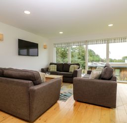 A living room with three gray sofas a wooden coffee table a flat screen tv on the wall and large windows overlooking trees at Maple in Truro