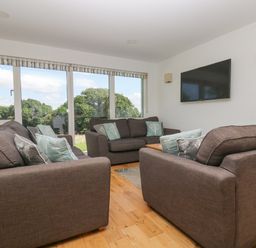 A living room with three sofas facing a wall mounted TV and large windows showing trees at Willow in Truro