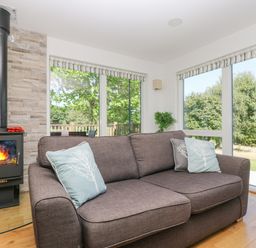 A living room with a gray sofa with pillows near a wood burning stove and large windows showing trees outside at Willow in Truro