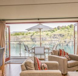 A living area with armchairs and dining chairs facing a balcony with table and umbrella overlooking a lake and rocky hillside at Boavista in Carnforth