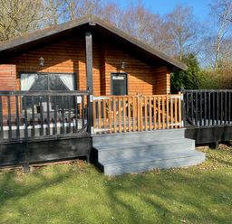 A wooden cabin with a porch and railing surrounded by trees and grass at Sunset Lodge in Tattershall