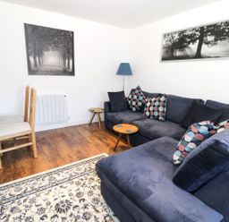 A living room with a blue sectional sofa with patterned cushions two wooden side tables a wooden dining table with chairs a floor lamp and black and white nature pictures on the walls at Skovhaven in Catton