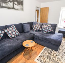A living room with a dark blue sectional sofa with patterned cushions a small wooden table a window with flowers on the sill and a patterned rug on the floor at Skovhaven in Catton