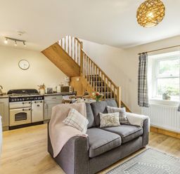 A kitchen and living area with a gray sofa under stairs next to a window with checked curtains at Briar Cottage in Edlingham
