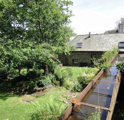 A garden with trees and plants next to a stone house with a water channel in the foreground at Water Barn in Manaton near Lustleigh