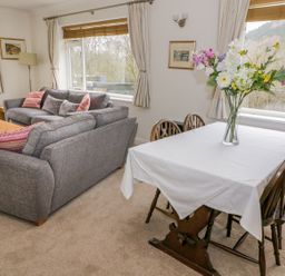 A dining table with a white tablecloth and a vase of flowers next to a living area with gray sofas in a room at The Dene in Betws-y-Coed