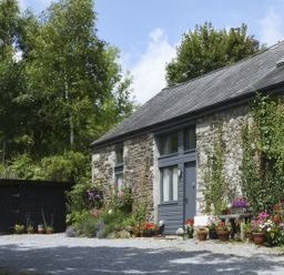 Stone cottage with gray door and windows surrounded by plants and flowers with gravel driveway at The Stone Barn Cottage in Holne