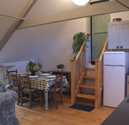 A dining area with a wooden table and chairs next to wooden stairs and a kitchen with a refrigerator at The Stone Barn Cottage in Holne
