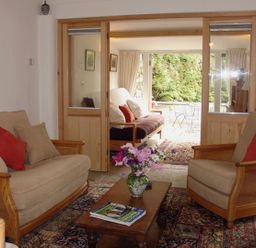 A living room with sofas and a coffee table with flowers and magazines separated by wooden framed glass doors from a sunroom with a sofa and outdoor furniture at Burham Barn in Dousland near Yelverton