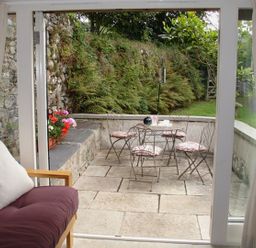 A patio with metal chairs and table with cushions viewed through sliding glass doors from a room with a wooden-framed sofa Burham Barn Dousland near Yelverton