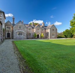 A large stone manor house with multiple chimneys and leaded windows facing a gravel path and a lawn with trees at Great Bidlake Manor in Bridestowe near Lydford
