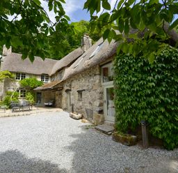 A stone cottage with a thatched roof and outdoor metal table and chairs surrounded by greenery at Bagtor Mill in Ilsington near Haytor Vale