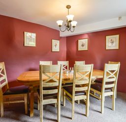 A dining room with a wooden table and eight chairs on carpet with framed pictures on red walls at Bagtor Mill in Ilsington near Haytor Vale