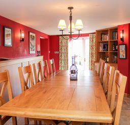 A dining room with a long wooden table surrounded by wooden chairs and a bookshelf against a red wall at Sandridge Barton in Stoke Gabriel