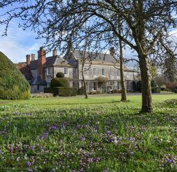 A large stone house with chimneys surrounded by trees and a grassy garden with purple and white flowers at Wootton House in Butleigh Wootton near Compton Dundon