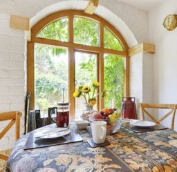 A dining area with a round table covered with a floral tablecloth near arched wooden windows at Pittards Farm Cottage in South Petherton