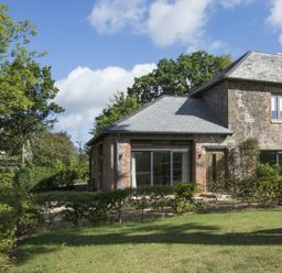 A stone house with a slate roof surrounded by bushes and trees with a wooden bench on the grass at Grooms Cottage Talaton near Ottery St Mary
