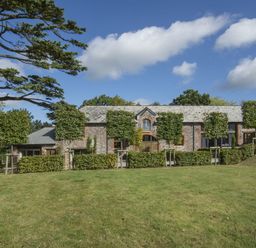 A stone building with a slate roof behind a row of pruned trees and hedges with a grassy lawn and a large tree on the left at Grooms Cottage Talaton near Ottery St Mary