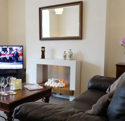 A living room with a brown leather sofa a wooden coffee table with wine glasses and a bottle of champagne a fireplace a TV showing a news program and a side table with pink flowers in a vase at 2 Carlton Mews in Exmouth