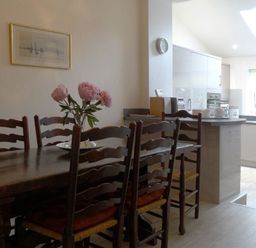 A dining area with a wooden table and chairs with cushions and a vase of flowers next to a kitchen with white cabinets and a countertop at 2 Carlton Mews in Exmouth