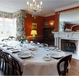 A dining room with a long table set for a meal chairs floral curtains a chandelier a fireplace and a large mirror at Abbots Manor Combe Raleigh near Honiton