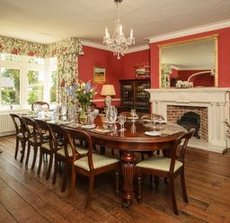 A dining room with a long wooden table set with glasses and cutlery a fireplace and floral curtains at Abbots Manor in Combe Raleigh near Honiton