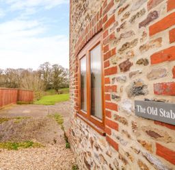 A stone and brick exterior wall with a window and a sign reading The Old Stable with a wooden fence and grassy area outside at The Old Stable in Sidbury near Sidmouth