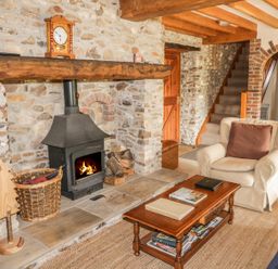A living area with a wood stove in a stone wall fireplace a wooden coffee table with books an armchair a wicker basket and stairs at Swallows Cottage in Honiton