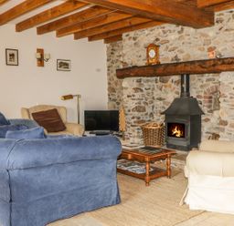 A living room with a blue sofa beige armchairs a wooden coffee table a stone fireplace with a wood stove and wooden beams on the ceiling at Swallows Cottage in Honiton