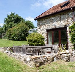 A stone house with a tiled roof and wooden doors next to a sunken patio with wooden benches and a table surrounded by a low stone wall in a grassy garden at Battens in Colyton